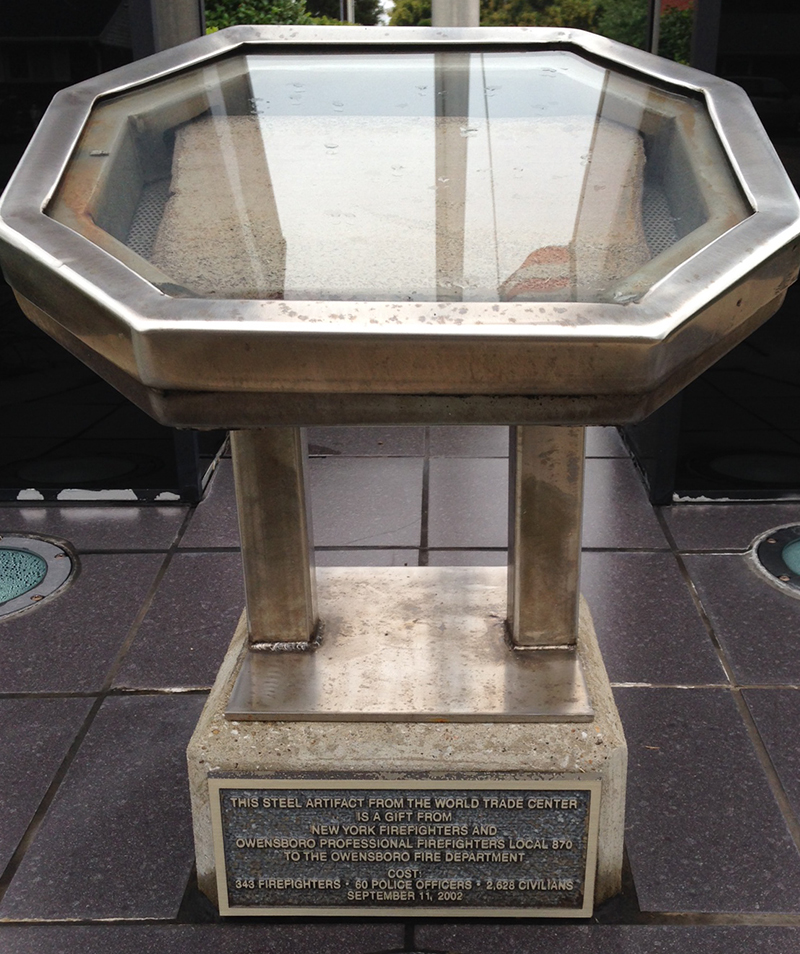 The plaque reads: "This steel artifact from the World Trade Center is a gift from New York Firefighters and Owensboro Professional Firefighters Local 870 to the Owensboro Fire Department. Cost: 343 Firefighters - 60 Police Officers - 2,628 Civilians. Septemner 11, 2002"
