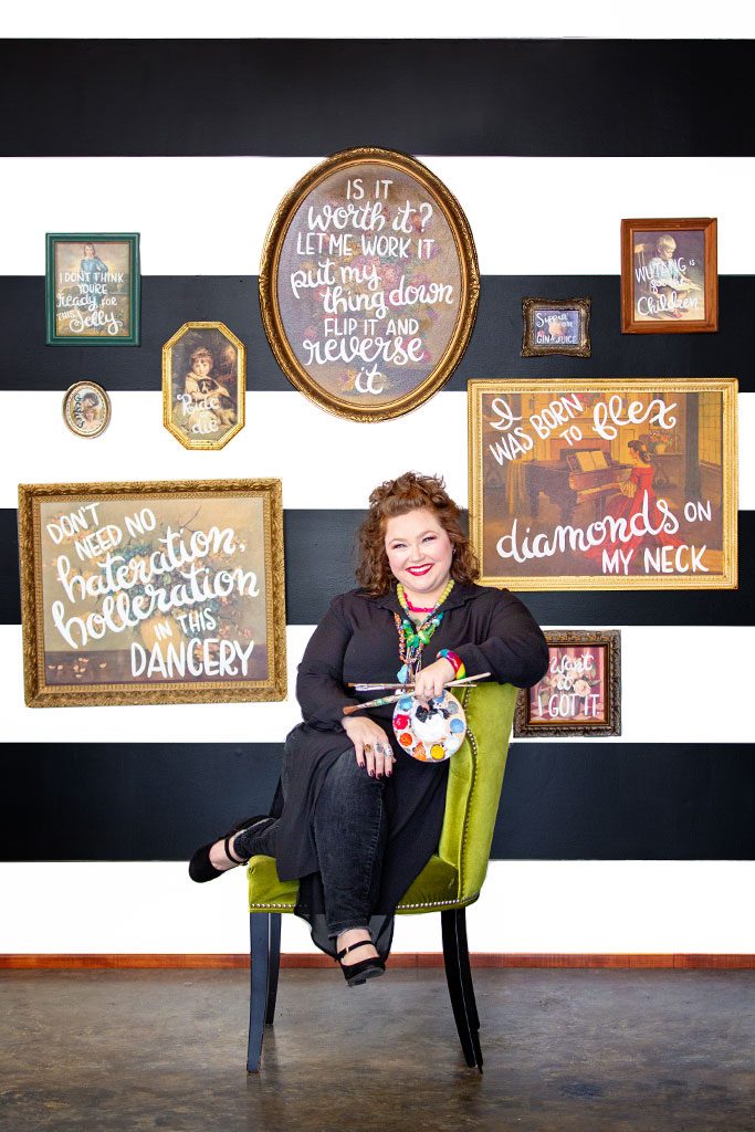 Maddox sits in a chair as her vintage paintings with bold, white, text hang on display behind her.