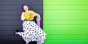 Mary Katherine Maddox, owner of M.porium, poses in a black and white polka dotted skirt, paint supplies in her hand, and a huge smile on her face.
